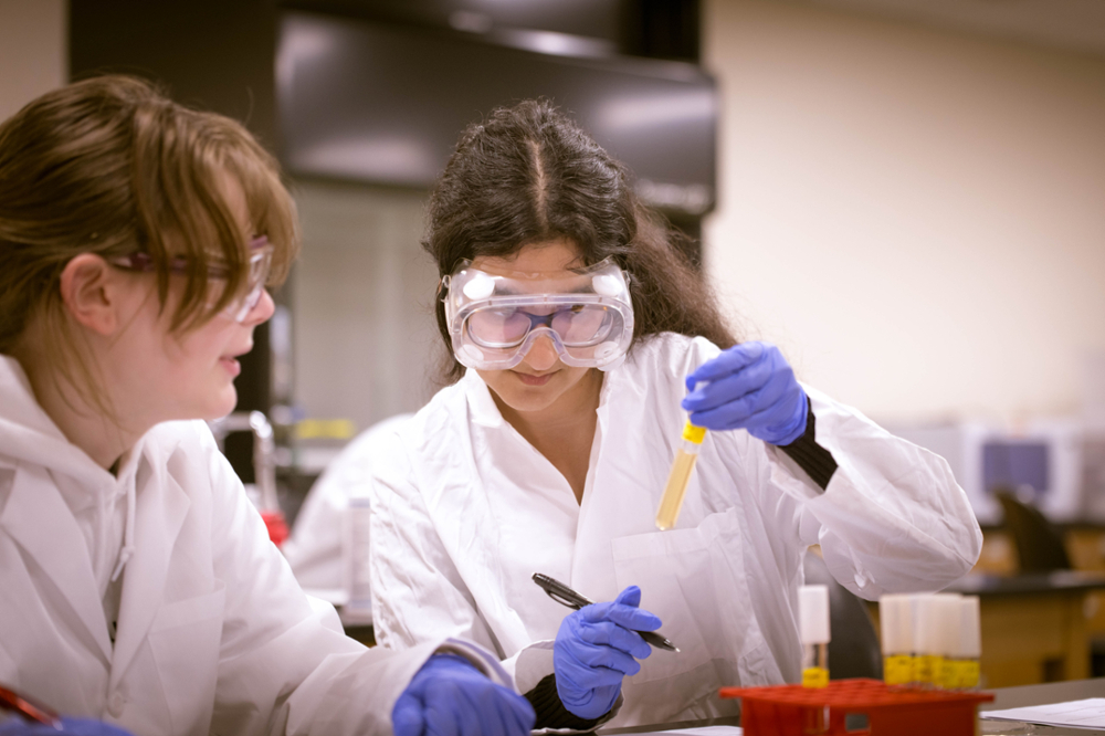 Two female students in white lab coats and safety goggles examine a yellow liquid specimen.