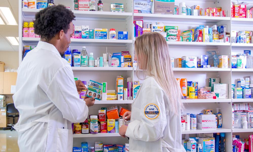 Pharmacist and WNE student study drug labels in front of shelf.