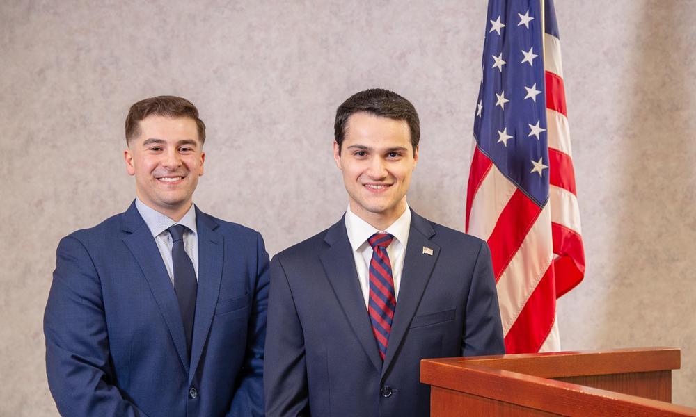 Two men in suits stand in front of the American flag