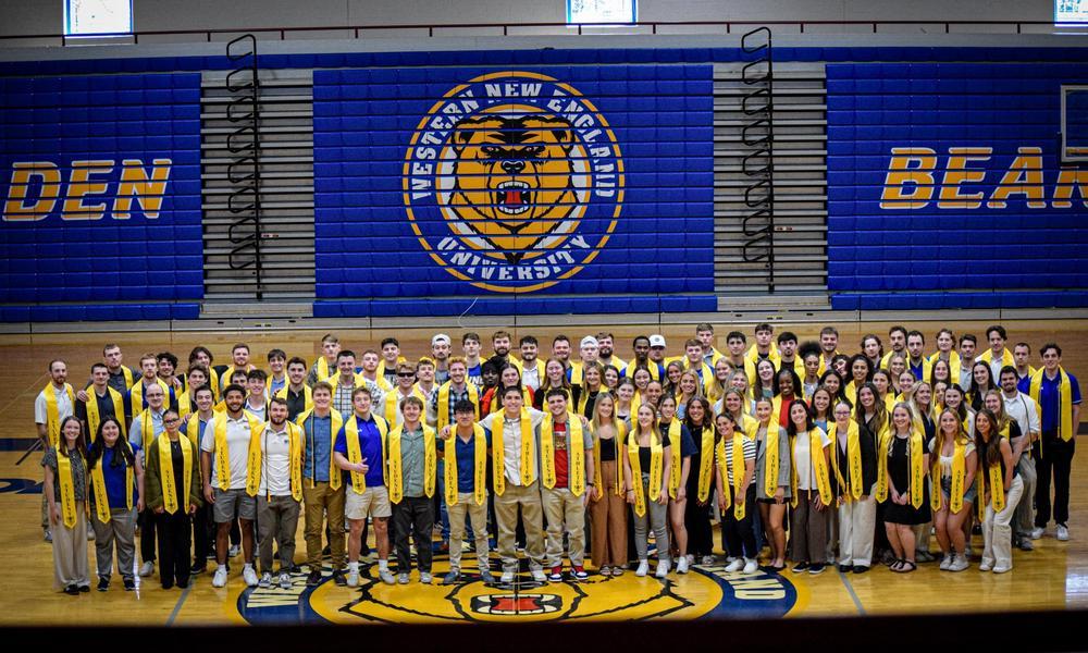 Shot of 56 WNE student athletes on gym floor under the Golden Bear logo.