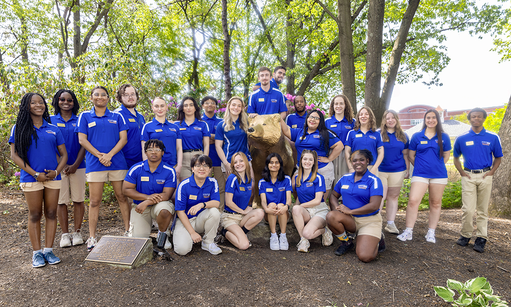 23 WNE students in blue WNE shirts pose around the Golden Bear