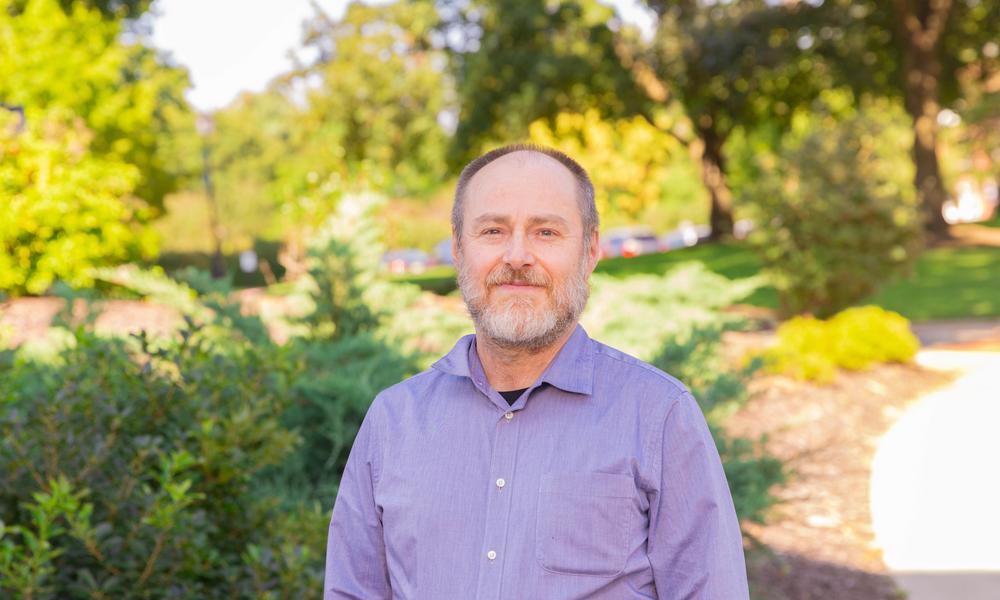 Professor Aydogdu stands outside in front of trees on a summer day.