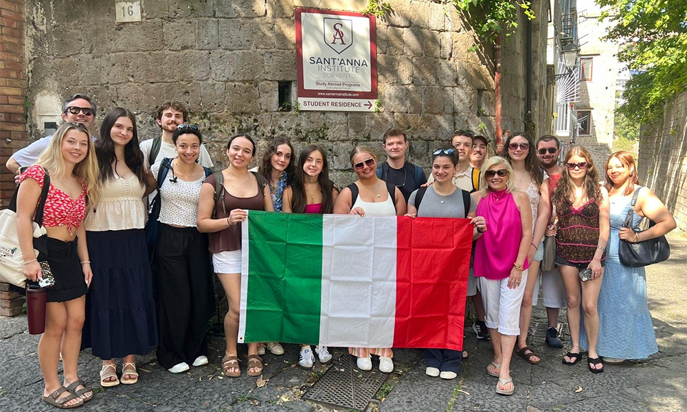 Sixteen students stand in front of an ancient stone wall holding an Italian flag.