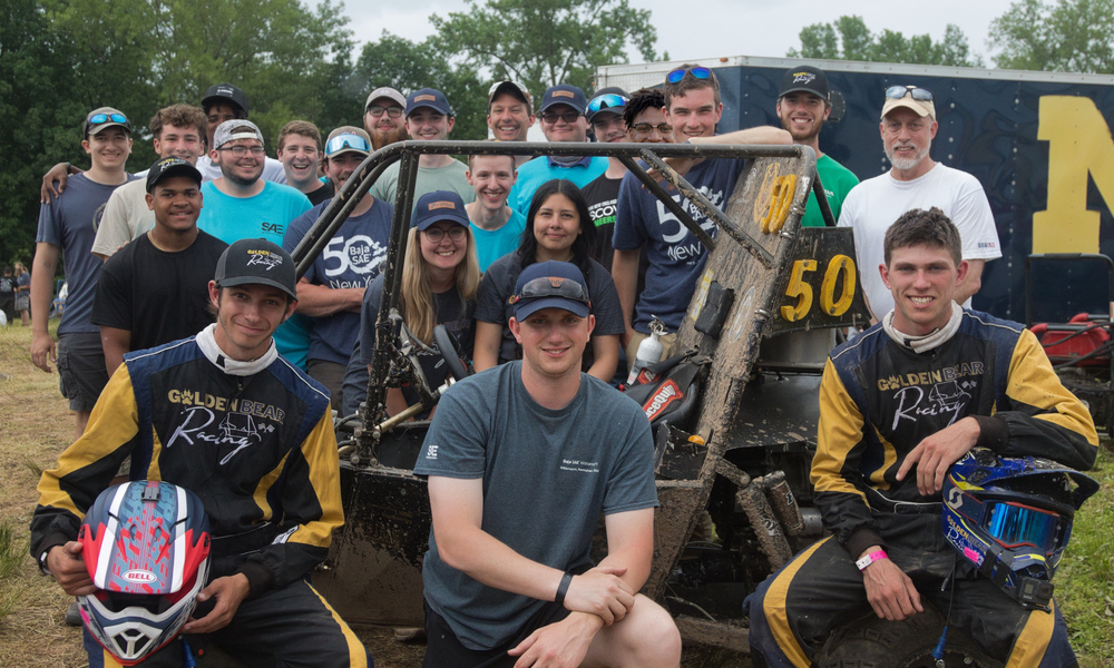 Approximately 30 students pose around a muddy baja car with big smiles.