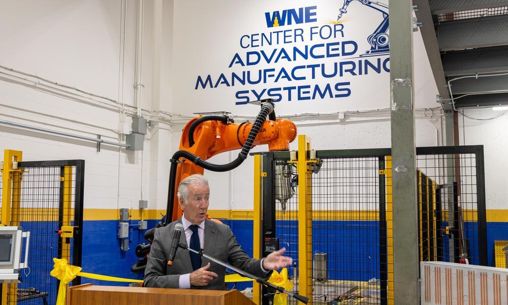 Congressman Richard Neal points while standing at podium in front of a giant orange robotic welding arm.