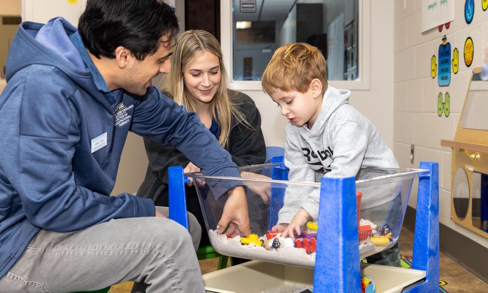 Two OTD students work with a pediatric OTB patient, a young boy of about 5 years old in a sand box.