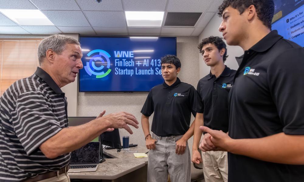 Man on left talks to three students in black polos about their fintech project with screen behind them announcing the Fintech + AI413 Start Up Launch series.