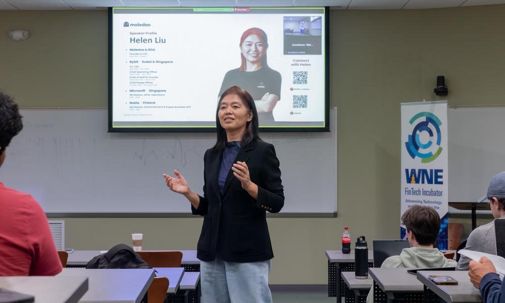 Helen Liu stands in front of a classroom of fintech students with a screen of her credentials behind her.