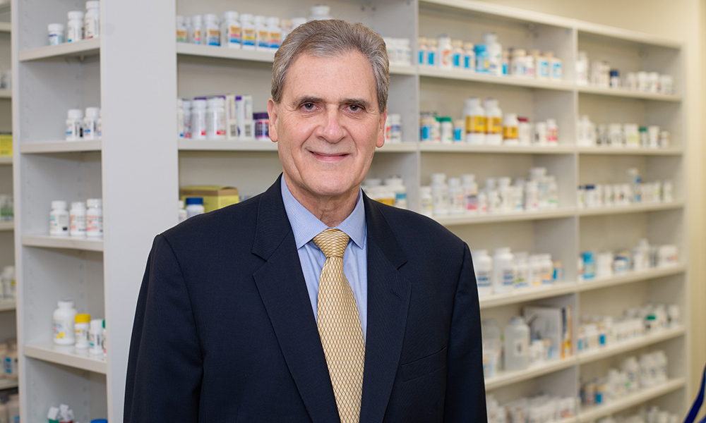 Head shot of researcher John Pezzuto in blue sport coast standing in front of shelves of medicine.